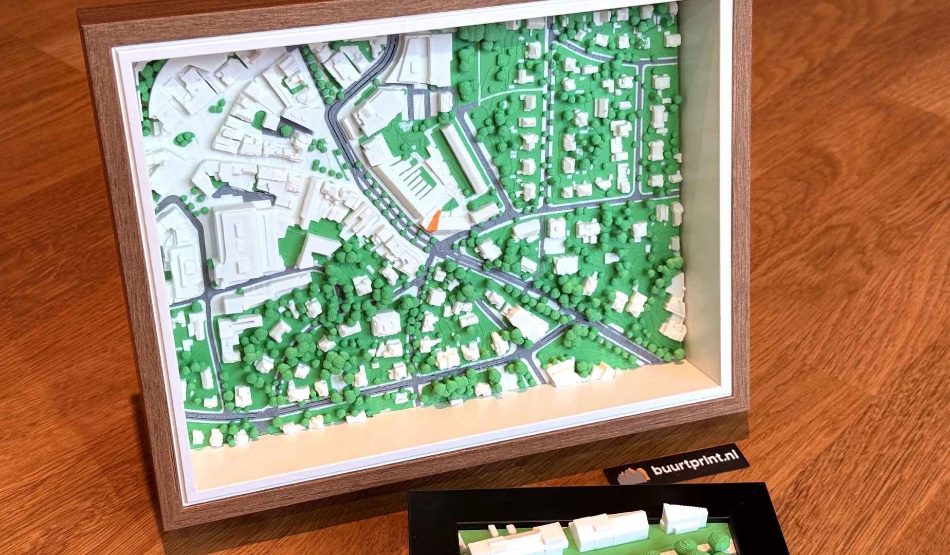 Geprinte maquette van een buurt in een houten lijst, van boven gezien, met witte huizen, groene bomen en het eigen huis in oranje